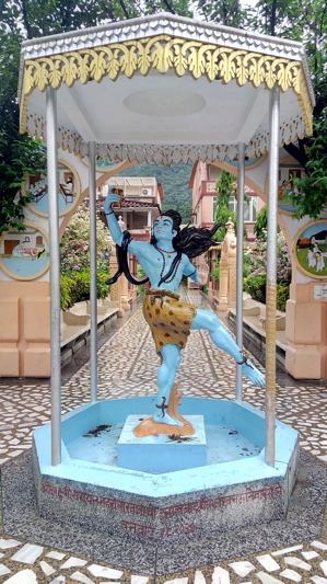 Statue of blue Lord Shiva with one leg up under umbrella in middle of buildings