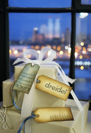 Stack of gifts in white paper with Hanukkah-themed tags, nighttime cityscape outside window in background