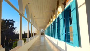 White walkway, open-air, with poles and blue shutters overlooking gardens below