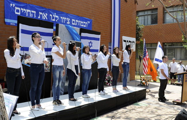Young people lined up on stage, outdoors, with blue banners and Israeli flags on building behind stage
